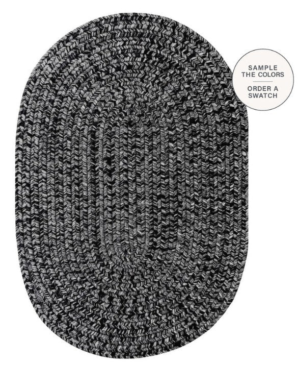 An oval braided area rug in a black and white heathered pattern, shown from a top-down view on a white background with a circular text overlay in the top right that reads 'SAMPLE THE COLORS ORDER A SWATCH'.