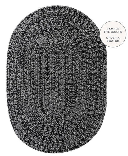 An oval braided area rug in a black and white heathered pattern, shown from a top-down view on a white background with a circular text overlay in the top right that reads 'SAMPLE THE COLORS ORDER A SWATCH'.