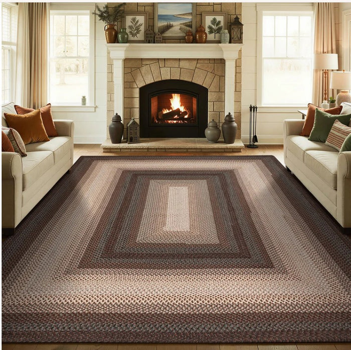 A large braided area rug with concentric rectangular patterns in shades of brown, beige, and gray, shown in a cozy living room setting with a fireplace, sofas, and windows.