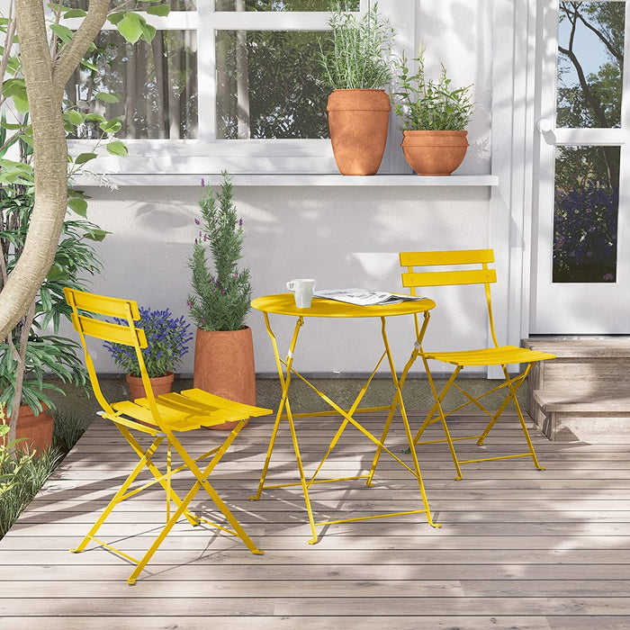 Yellow metal chairs and table set on a wooden deck with plants and a white wall in the background.