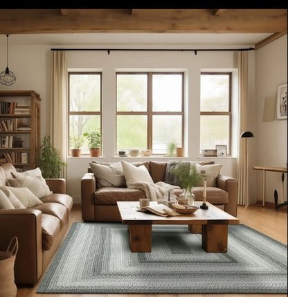 A styled living room interior with a large rectangular area rug in muted blue-gray tones, a brown leather sofa, a matching coffee table, wood ceiling beams, and three tall windows with beige curtains.