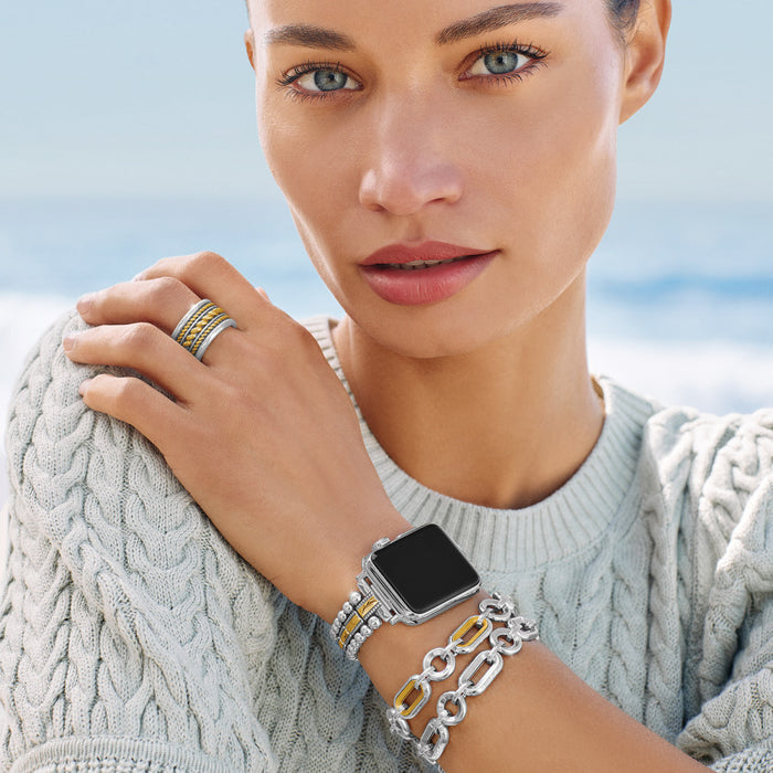 A woman wearing the Medici Two Tone Link Bracelet poses against a blurry coastal background. She is showcasing a smartwatch band, bracelets, and a ring on her left hand.