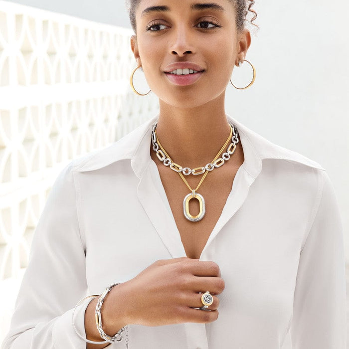 A woman is wearing the Medici Two Tone Link Bracelet with matching earrings, necklaces, and a ring, against a white blouse and patterned background. She is looking upward with a soft smile.
