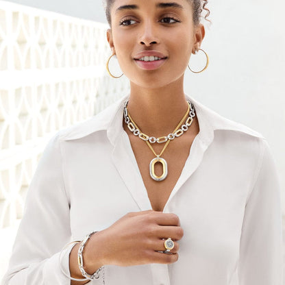 A woman is wearing the Medici Two Tone Link Bracelet with matching earrings, necklaces, and a ring, against a white blouse and patterned background. She is looking upward with a soft smile.