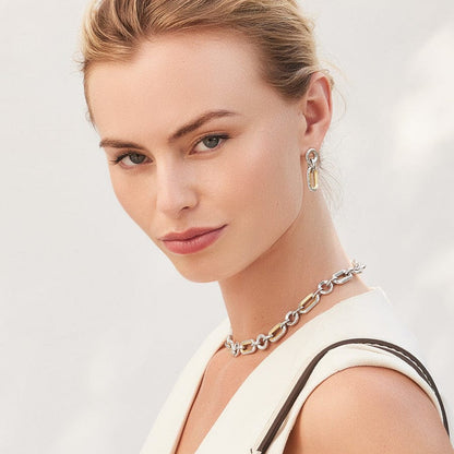 A woman her wearing Medici Two Tone Link Post Drop Earrings and a chunky chain necklace. She appears poised against a soft, neutral background.