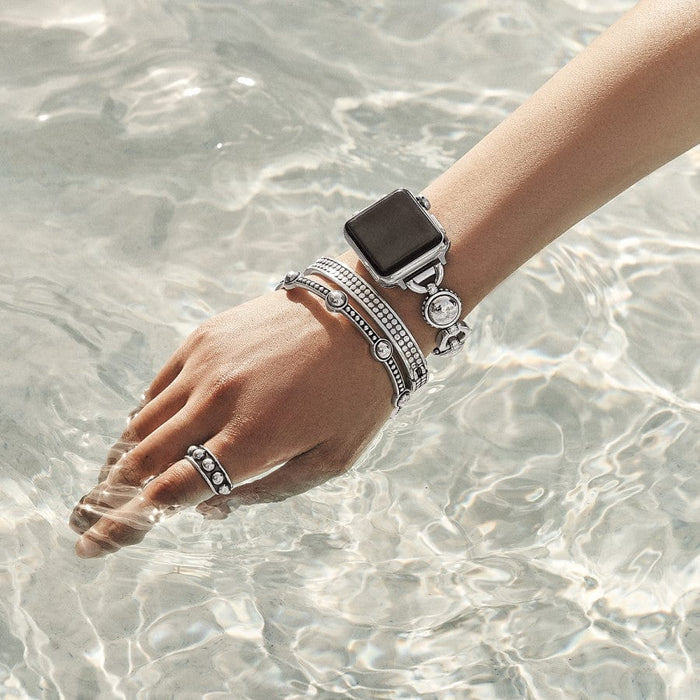 A hand adorned with a Pretty Tough Bold Apple Watch Band, rings, and bracelets extends into clear, rippling water under bright light. The jewelry includes a mix of metallic and bejeweled designs.