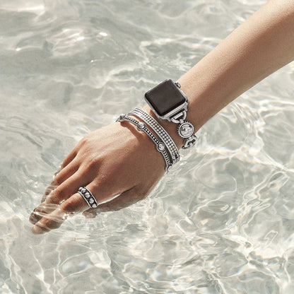 A hand adorned with a Pretty Tough Bold Apple Watch Band, rings, and bracelets extends into clear, rippling water under bright light. The jewelry includes a mix of metallic and bejeweled designs.